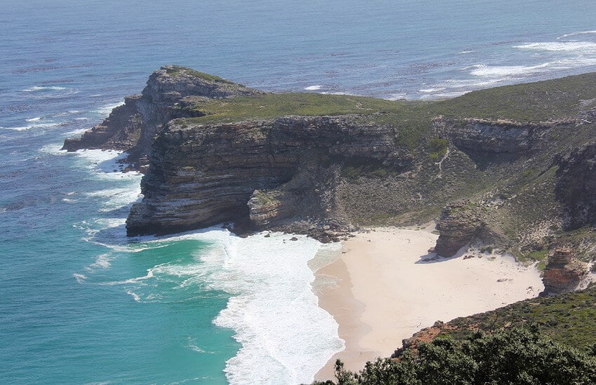 Eine hohe steile Felsklippe, dazwischen ein kleiner flacher Sandstrand.