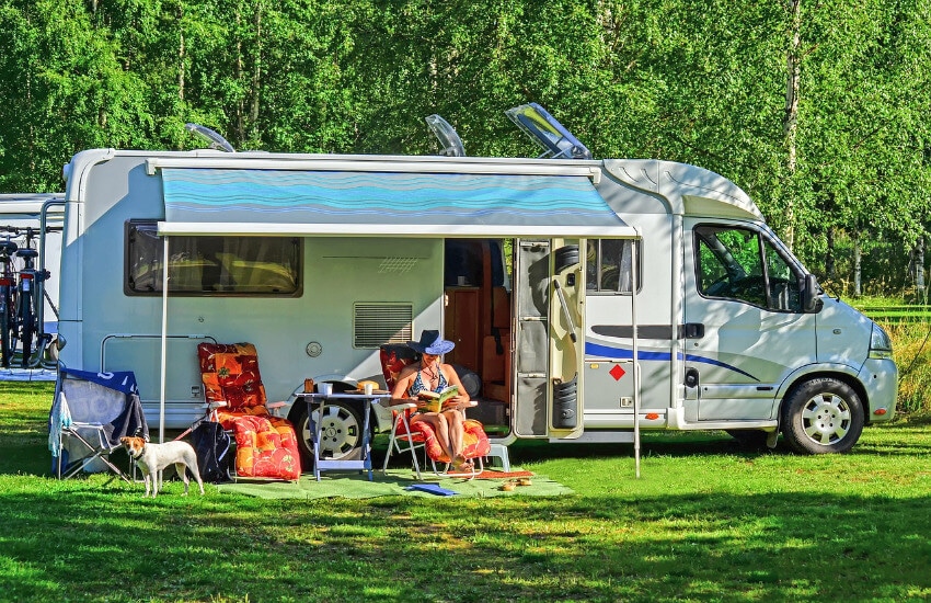 Ein Wohnmobil parkt an einem Campingplatz mit einem kleinen Vordach. Davor eine Frau im Stuhl und list ein Buch.