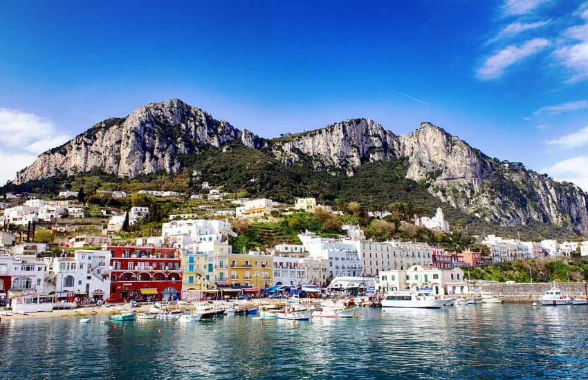 Einige Schiffe ankern am Hafen dahinter eine Häuserreihe eng zusammengebaut und im Hintergrund die Berge von Capri.