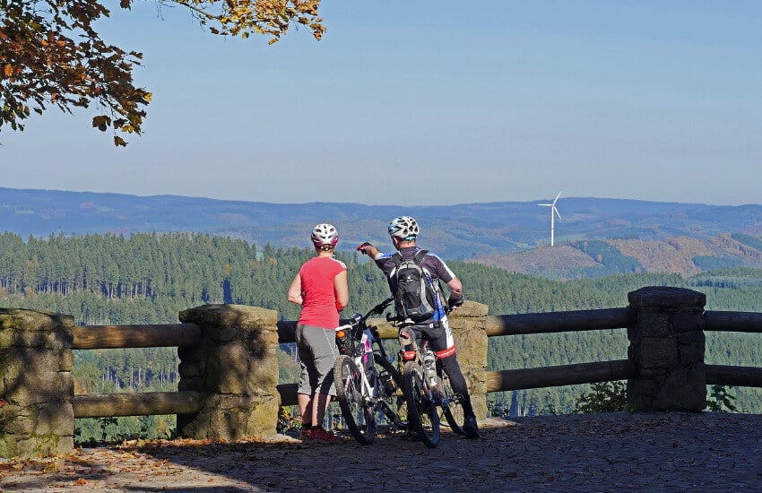 Zwei Radfahrer blicken vom Berg Hohe Bracht Richtung einer mit Wald bedecken Landschaft.