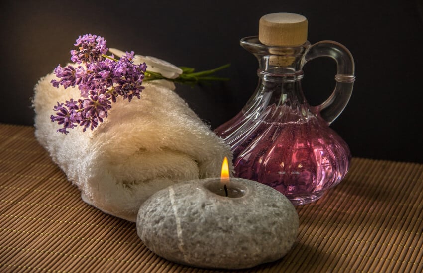 Accessoires für eine Wellnessbehandlung. Ein gerolltes Handtuch mit einer Lavendelblüte oben drauf, ein Glas mit Lavendelöl und ein Stein mit einer Kerze.