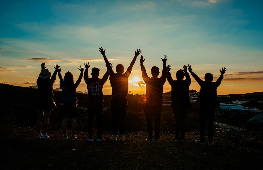 Klassenfahrt Eine Gruppe fröhlicher Jugendlicher jubelt mit erhobenen Armen vor einem Sonnenuntergang.