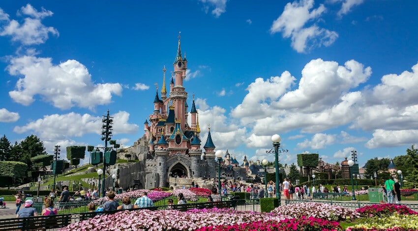 Disneyland Paris Das rosa Dornröschenschloss im Disneyland Paris mit blauen Türmen, umgeben von einer Menschenmenge an einem sonnigen Tag.