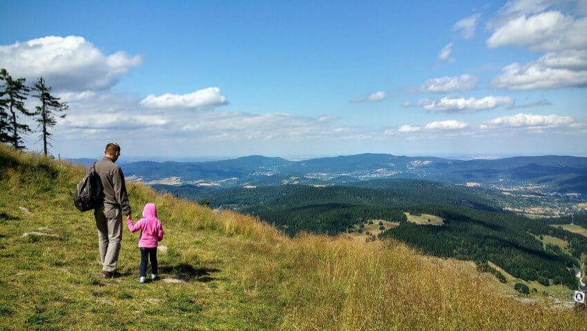 Familienurlaub mit Kleinkind Ein Vater hält sein kleines Kind an der Hand und blickt vom Berg aus über eine hügelige, mit Bäumen bewachsene Landschaft in Österreich.